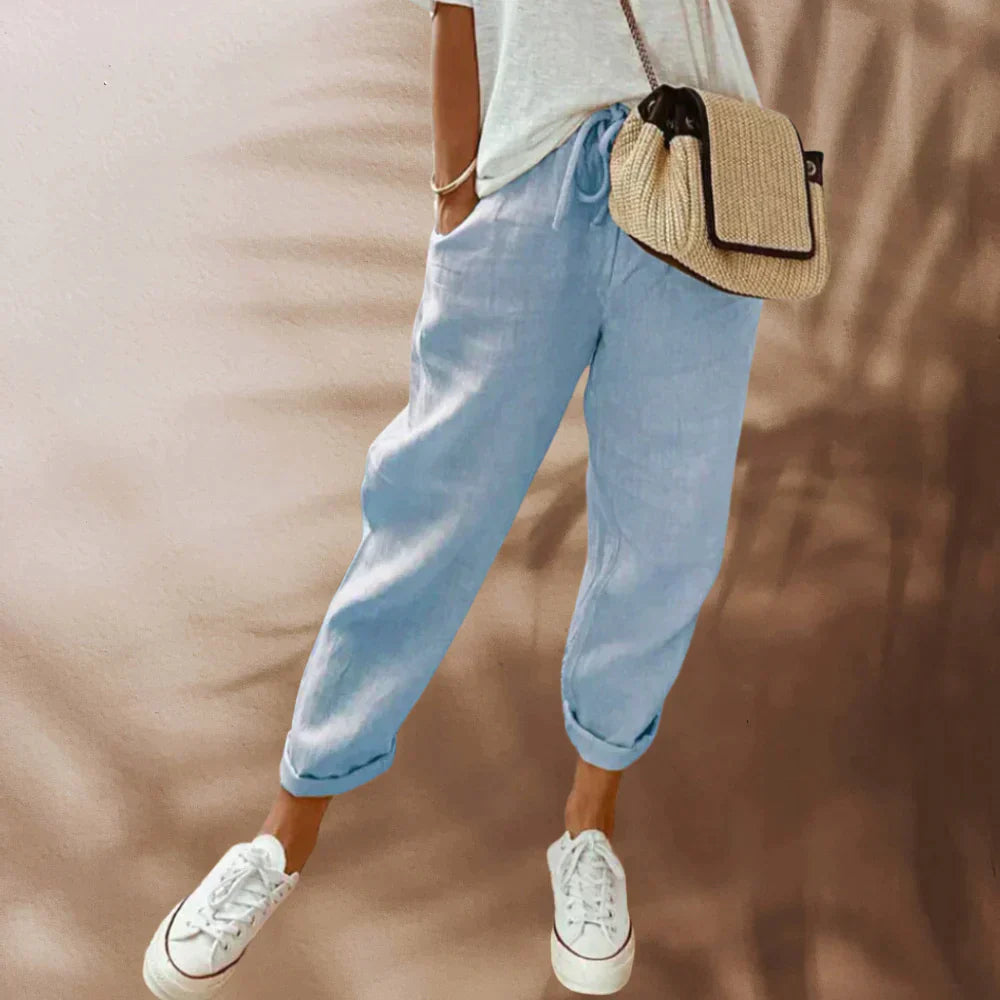 Pantalon en lin bleu clair pour femme, style décontracté, avec poches, associé à un t-shirt blanc, sac en osier et baskets blanches.