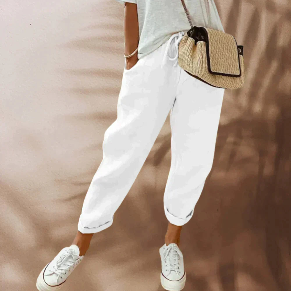 Femme portant un pantalon blanc décontracté, t-shirt gris, baskets blanches et sac à bandoulière en paille. Mode estivale chic et décontractée.