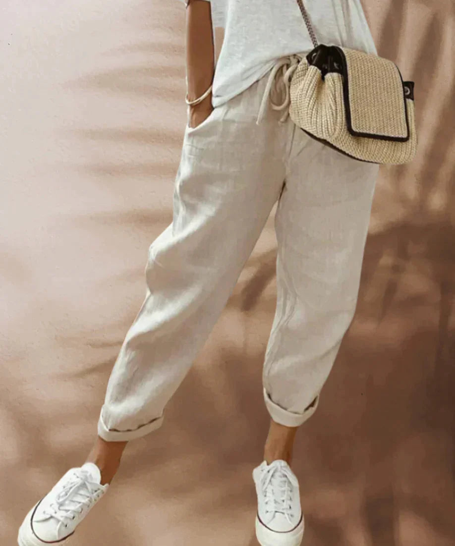 Personne portant un pantalon en lin beige, t-shirt blanc, baskets blanches et sac en osier. Mode décontractée, style estival, tenue confortable.