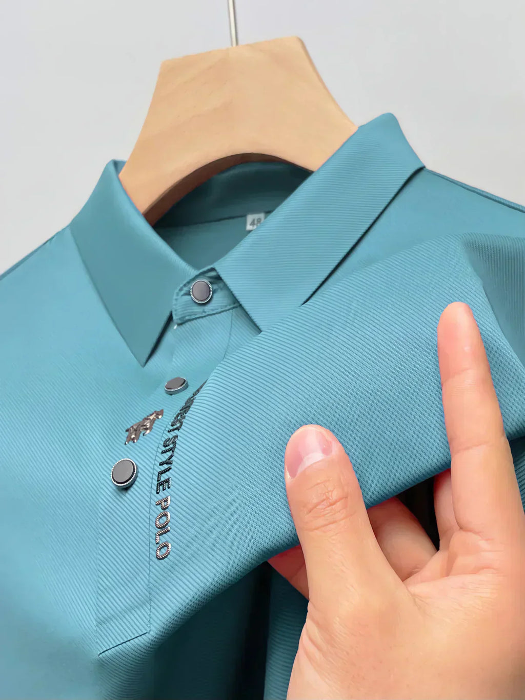 Polo bleu turquoise en tissu texturé, avec col boutonné et logo brodé "Gentle Polo", suspendu sur un cintre en bois.