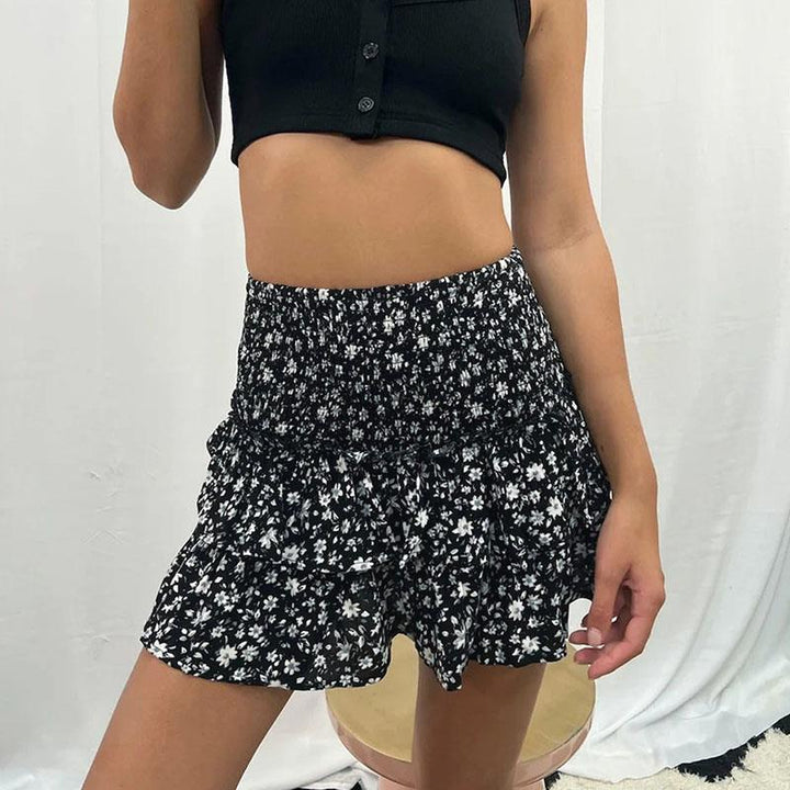 Jeune femme portant une jupe noire à motifs floraux blancs et un haut court noir, posant devant un fond blanc. Mode féminine, style estival.