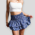 Femme en top blanc et jupe bleue à motifs floraux, posant sur fond gris. Mode estivale, tenue décontractée, style tendance.