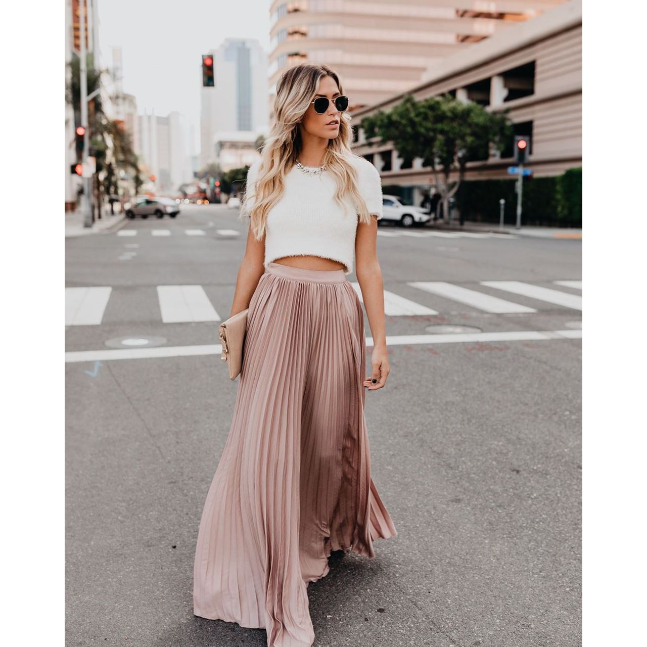Femme élégante en jupe plissée rose et haut blanc, marchant en ville. Mode urbaine chic, style décontracté, lunettes de soleil, sac à main beige.