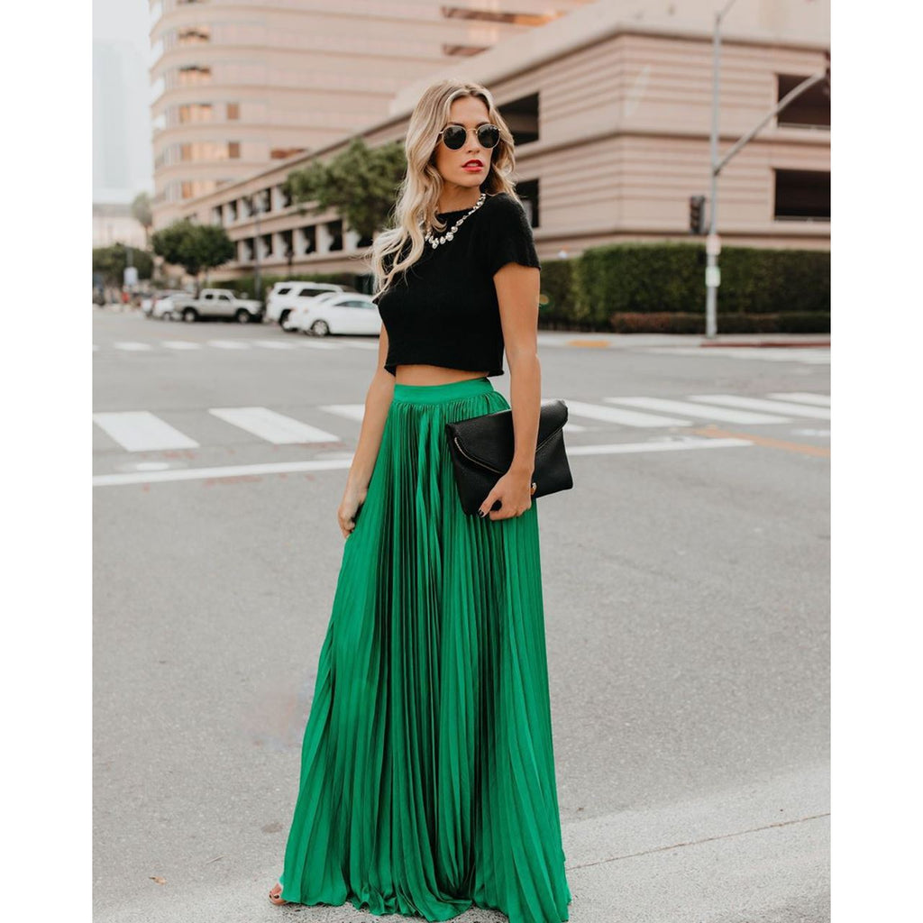 Femme élégante en jupe longue verte et haut noir, tenant une pochette noire, traverse une rue urbaine. Mode chic, style urbain, tenue tendance.