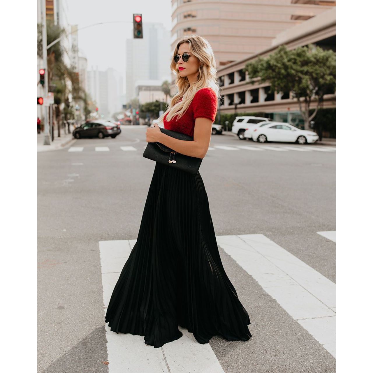 Femme élégante en jupe longue noire et haut rouge, tenant un sac à main noir, traverse une rue urbaine. Mode chic, style urbain, tenue tendance.
