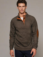 Homme portant un pull marron à col montant avec boutons et coudières en cuir, sur fond neutre. Mode masculine élégante et décontractée.