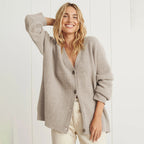 Femme souriante portant un cardigan beige en laine, manches longues, devant un mur blanc. Mode automne-hiver, style décontracté et élégant.