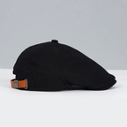 Casquette plate noire en tissu avec sangle réglable en cuir marron, posée sur une surface blanche, fond gris. Accessoire mode élégant et classique.