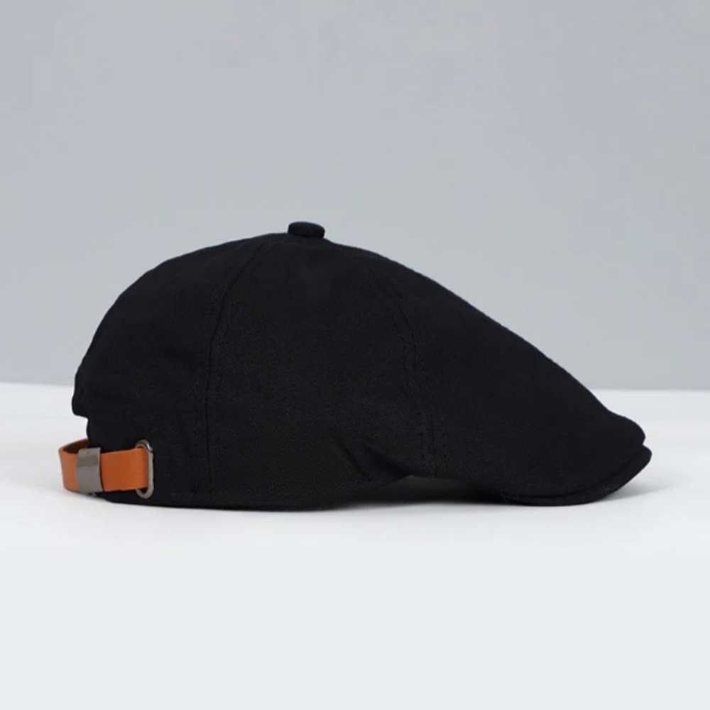 Casquette plate noire en tissu avec sangle réglable en cuir marron, posée sur une surface blanche, fond gris. Accessoire mode élégant et classique.