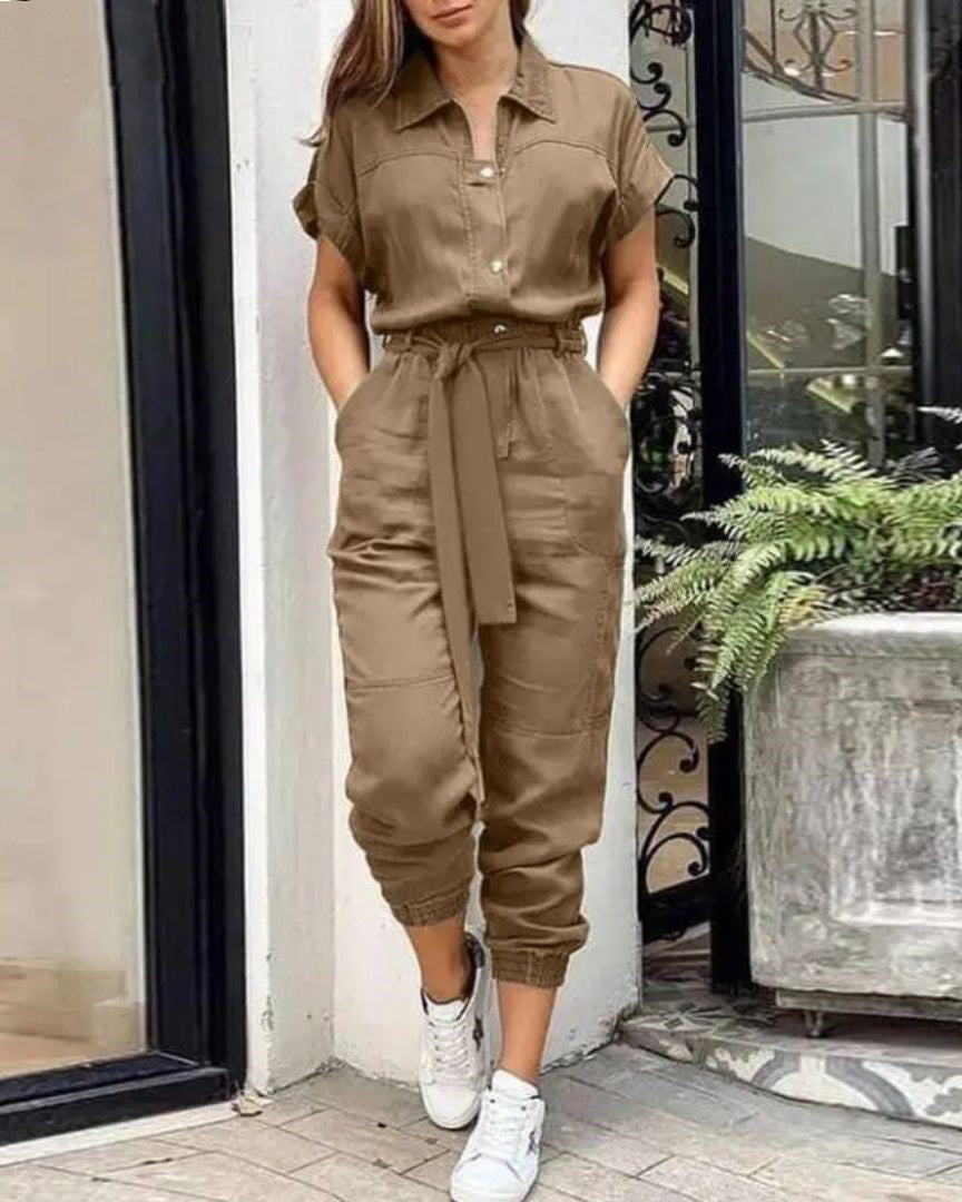 Combinaison marron élégante pour femme, manches courtes, ceinture nouée, portée avec des baskets blanches, devant une entrée moderne avec plantes.