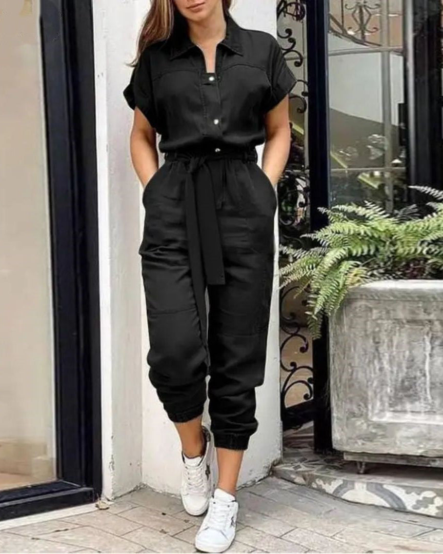 Femme portant une combinaison noire élégante avec ceinture, manches courtes, posant près d'une plante en pot, baskets blanches. Mode urbaine chic.