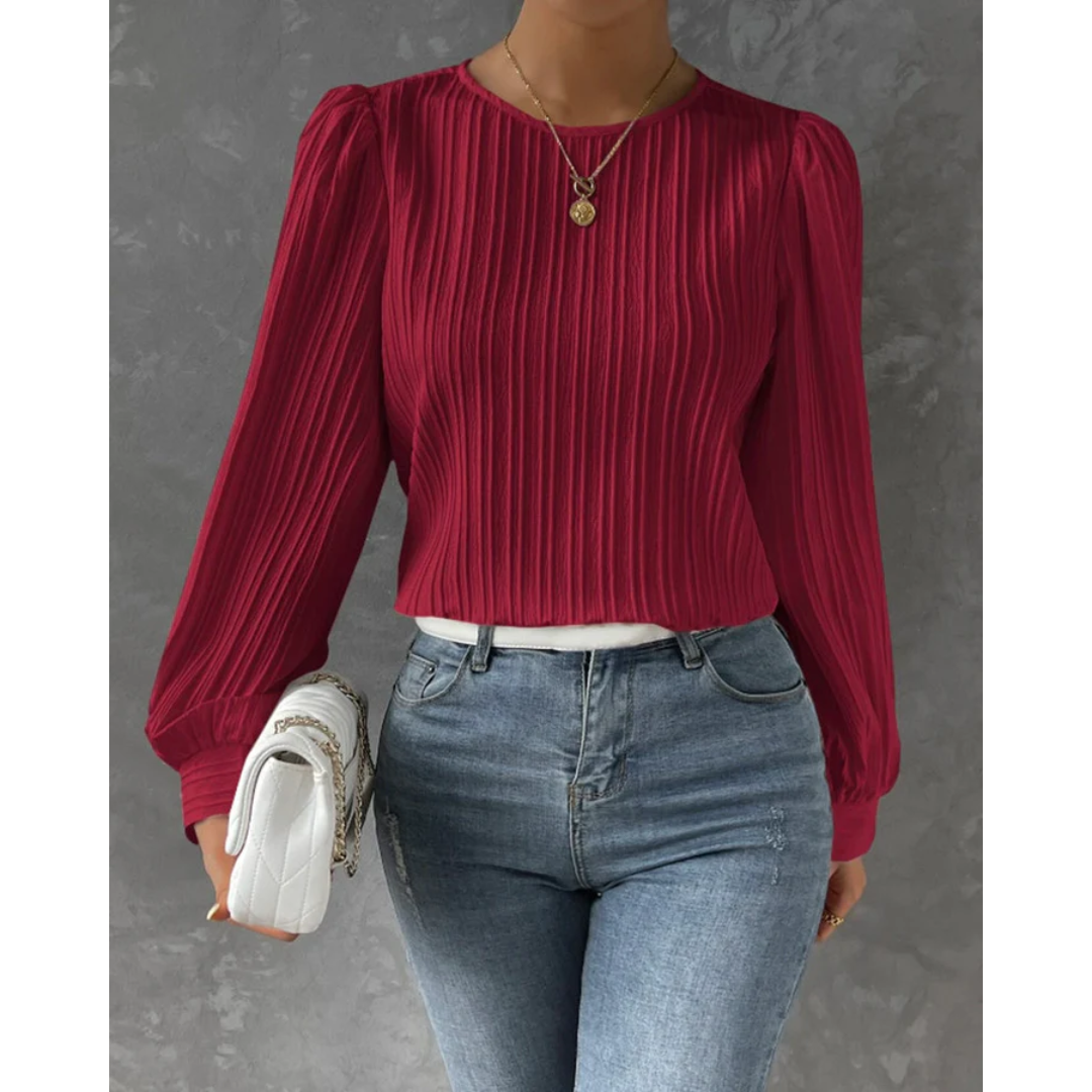 Femme portant un pull rouge à manches longues plissées, jean bleu ajusté, collier doré, et sac blanc. Mode féminine élégante et tendance.