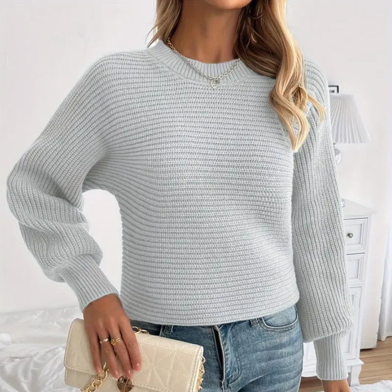 Femme portant un pull en tricot gris clair, manches longues, avec un sac à main beige. Mode automne, style décontracté, intérieur lumineux.