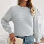 Femme portant un pull en tricot gris clair, manches longues, avec un sac à main beige. Mode automne, style décontracté, intérieur lumineux.