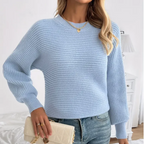 Femme portant un pull bleu clair en tricot, jeans décontracté, tenant un sac à main beige. Mode automne, style décontracté chic, accessoires dorés.
