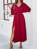Robe longue rouge élégante avec ceinture nouée, manches chauve-souris, fente latérale, portée par une femme devant une fenêtre. Mode féminine chic.