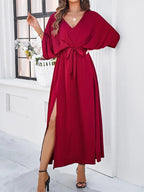 Robe longue rouge élégante avec ceinture nouée, manches chauve-souris, fente latérale, portée par une femme devant une fenêtre. Mode féminine chic.