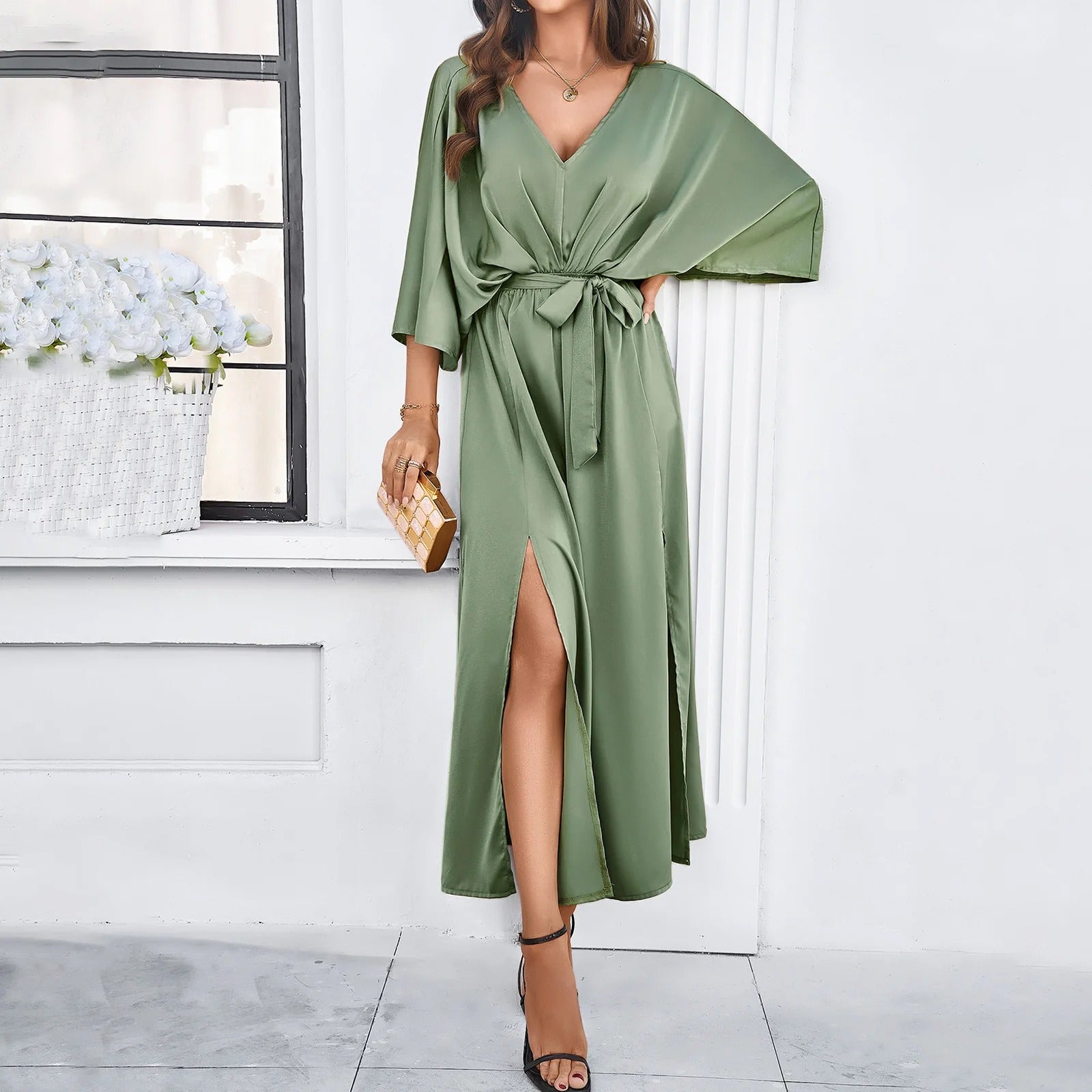 Robe longue verte élégante avec ceinture, manches larges et fente, portée par une femme tenant une pochette dorée, devant une fenêtre.