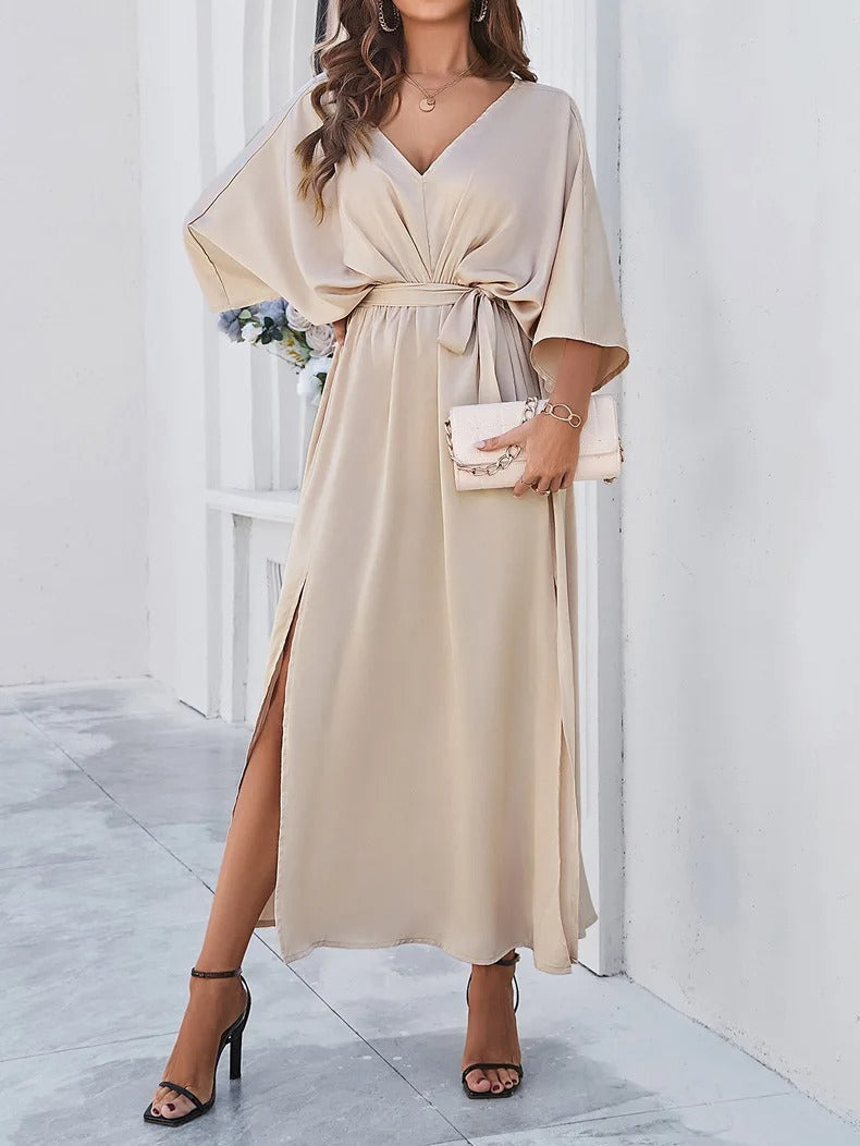 Robe longue beige élégante avec manches chauve-souris, ceinture nouée à la taille, portée avec des sandales à talons et pochette assortie.