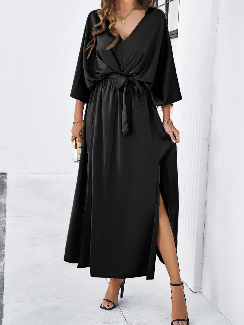 Robe longue noire élégante avec ceinture, manches chauve-souris et fente latérale, portée par une femme tenant un sac doré. Mode chic et sophistiquée.