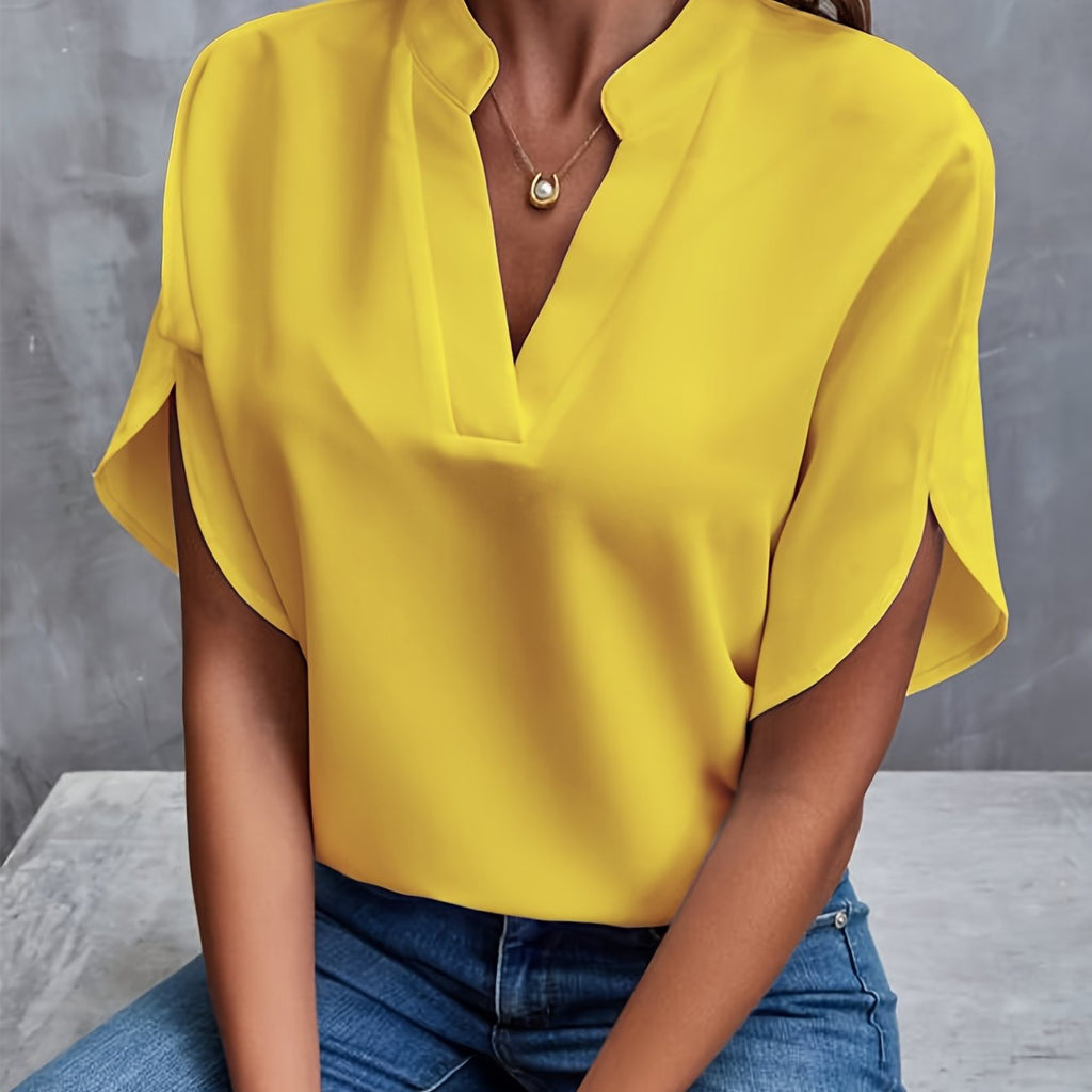 Femme portant un chemisier jaune vif avec un col en V, manches courtes et un collier doré, assise sur un fond gris. Mode féminine élégante.