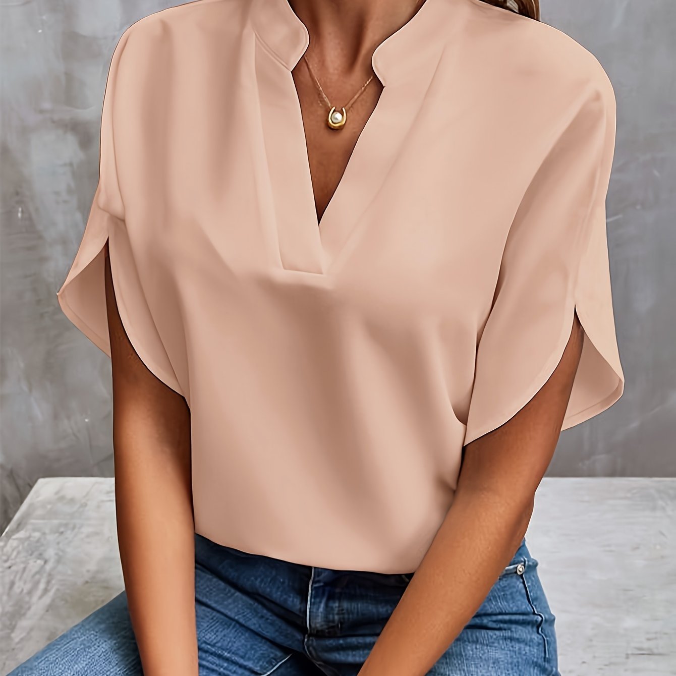 Blouse beige élégante avec col en V, manches courtes, portée par une femme assise, associée à un collier doré et un jean bleu. Mode féminine chic.