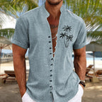 Homme en chemise en lin bleu avec motif palmiers, manches courtes, col mao, sur une plage tropicale avec parasols et mer en arrière-plan.