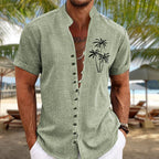 Homme portant une chemise en lin vert avec motif palmiers, sur une plage tropicale. Mode estivale, vacances, style décontracté.