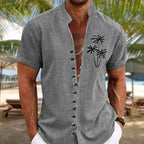 Homme portant une chemise grise à manches courtes avec motif de palmiers, sur une plage tropicale. Mode estivale, tenue décontractée, vacances.