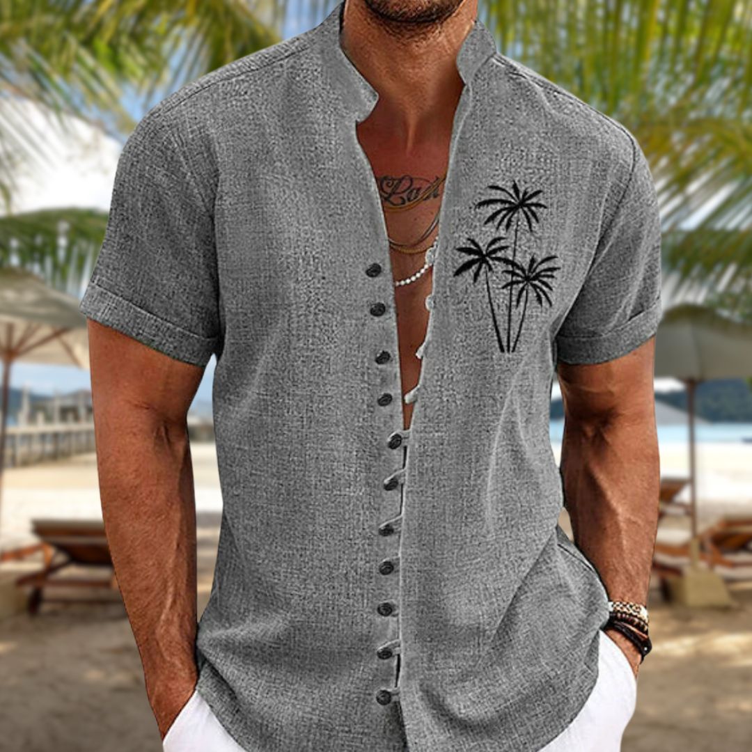 Homme portant une chemise grise à manches courtes avec motif de palmiers, sur une plage tropicale. Mode estivale, tenue décontractée, vacances.