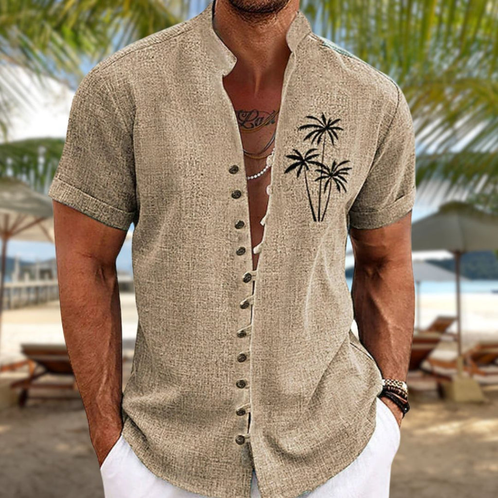 Homme portant une chemise en lin beige avec motif de palmiers, sur une plage tropicale. Mode estivale, vacances, style décontracté.