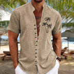 Homme portant une chemise en lin beige avec motif de palmiers, sur une plage tropicale. Mode estivale, vacances, style décontracté.