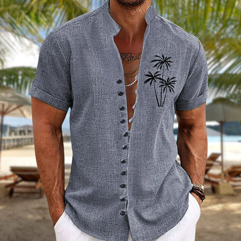 Homme portant une chemise grise à col mao avec motif de palmiers, sur une plage tropicale. Mode estivale, tenue décontractée, vacances bord de mer.