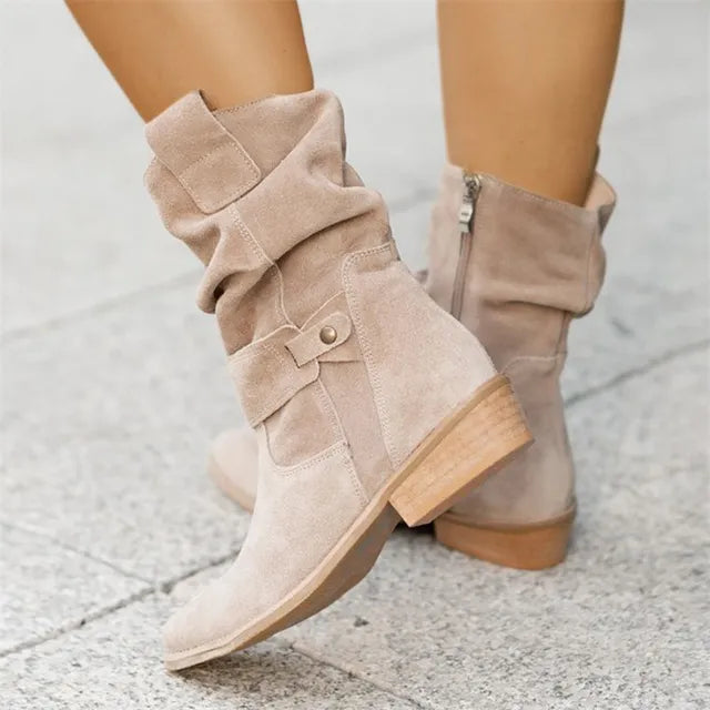 Bottes en daim beige pour femme avec talon bas, fermeture éclair latérale et design plissé, sur fond de trottoir gris. Mode automne-hiver.