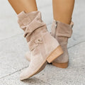Bottes en daim beige pour femme avec talon bas, fermeture éclair latérale et design plissé, sur fond de trottoir gris. Mode automne-hiver.