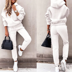 Femme en survêtement blanc, baskets blanches, sac à main noir, posant contre un mur gris. Mode décontractée, style urbain, tendance automne.