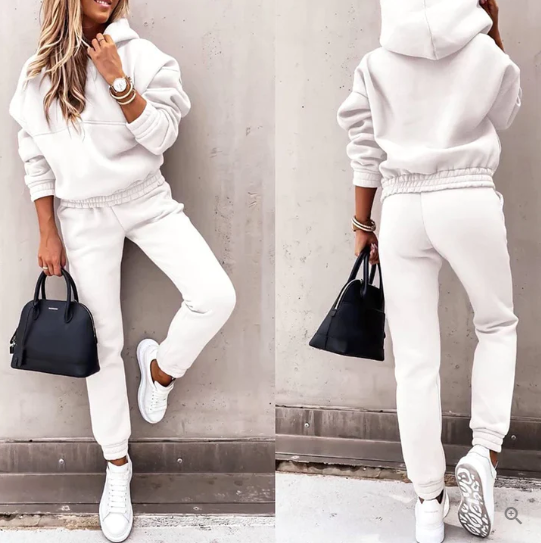 Femme en survêtement blanc, baskets blanches, sac à main noir, posant contre un mur gris. Mode décontractée, style urbain, tendance automne.