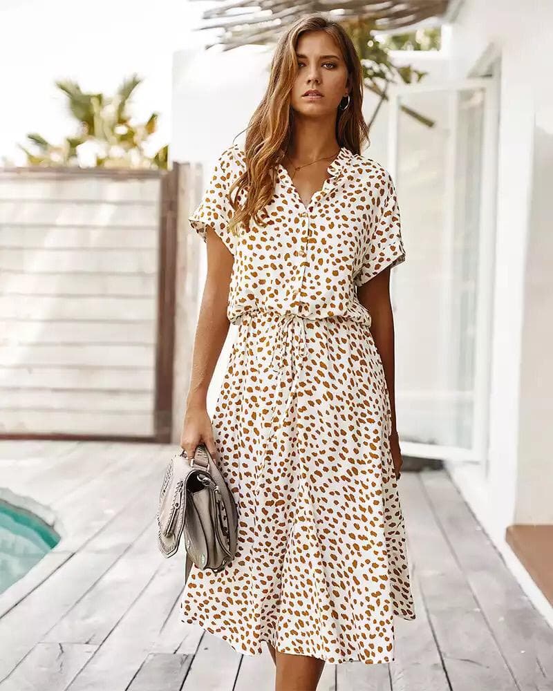 Femme portant une robe d'été à motifs léopard, debout près d'une piscine, tenant un sac à main gris. Mode estivale, style décontracté chic.