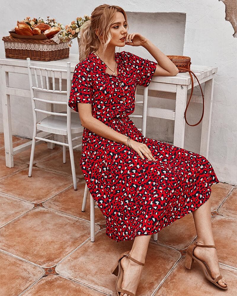 Femme assise en robe rouge à motifs léopard, sur une terrasse ensoleillée, avec des chaises blanches et des fleurs en arrière-plan. Mode estivale élégante.