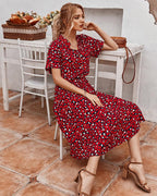 Femme assise en robe rouge à motifs léopard, sur une terrasse ensoleillée, avec des chaises blanches et des fleurs en arrière-plan. Mode estivale élégante.