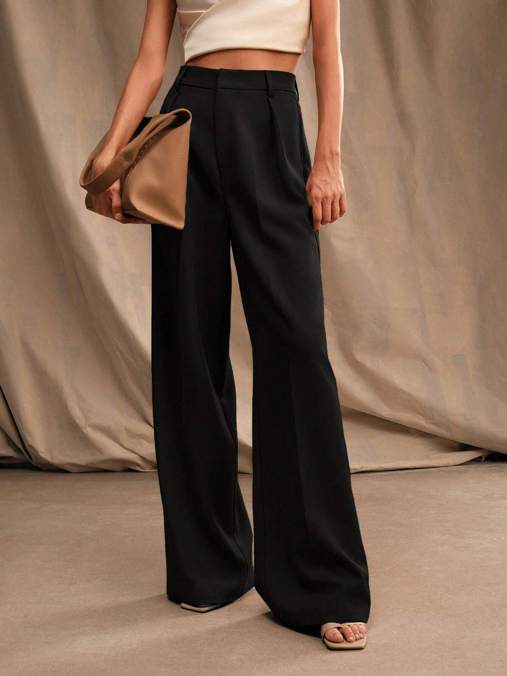 Femme portant un pantalon noir large et un haut blanc, tenant un sac à main beige. Mode élégante, style minimaliste, tenue chic.