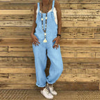 Femme portant une salopette en jean bleu clair, t-shirt blanc, collier long, baskets blanches, posant devant un mur en bois. Mode décontractée tendance.