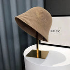 Chapeau cloche en maille beige sur support doré, devant une boîte Gucci. Accessoire de mode élégant et tendance.