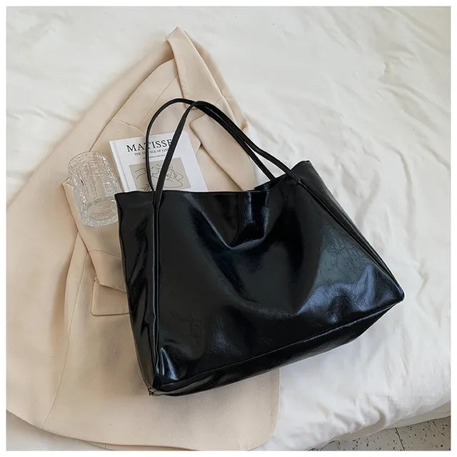 Sac à main noir en cuir brillant sur un lit blanc, accompagné d'un magazine et d'un manteau beige. Accessoire mode élégant et moderne.