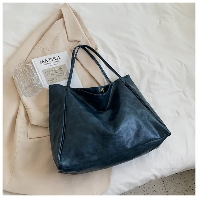 Sac à main en cuir noir élégant posé sur un manteau beige, avec un verre transparent et un livre d'art Matisse, sur un lit blanc. Accessoire mode chic.