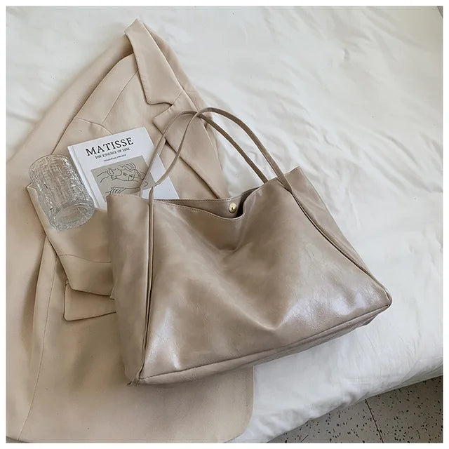 Sac à main en cuir beige sur un manteau crème, avec un livre "Matisse" et un verre en cristal, posés sur un lit blanc. Accessoires élégants.