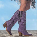 Bottes violettes en daim avec franges et talons blocs, portées par une femme sur fond de ciel bleu. Mode féminine, chaussures tendance.