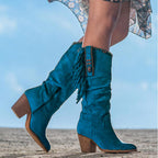 Bottes western bleues en daim avec franges et talons carrés, portées par une femme sur fond de ciel bleu. Mode féminine, style cowboy chic.