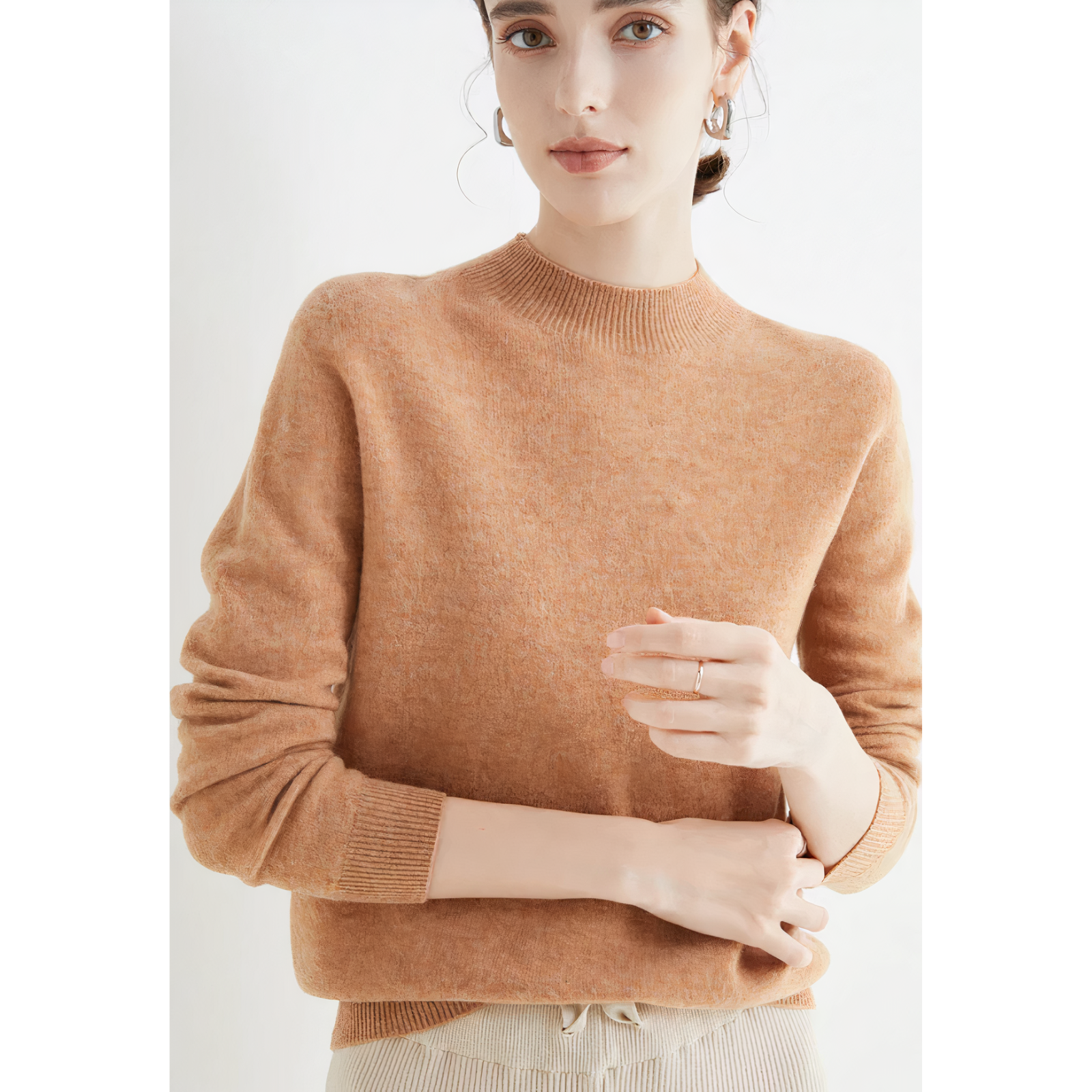 Femme portant un pull en laine beige, manches longues, col rond, posant contre un fond blanc. Mode automne-hiver, style élégant et décontracté.