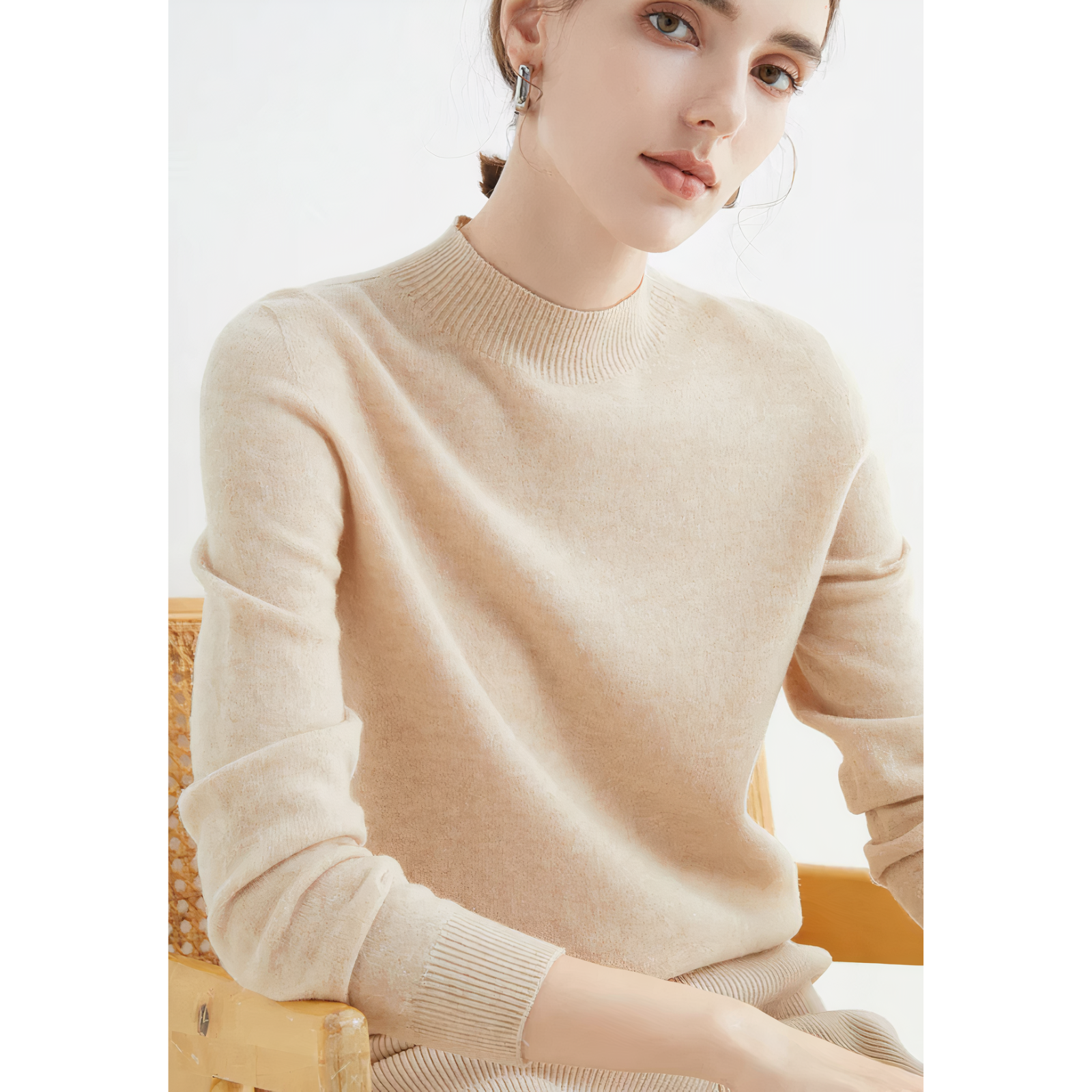 Femme portant un pull beige en laine, assise sur une chaise en bois. Mode automne-hiver, style élégant et minimaliste. Accessoire: boucles d'oreilles.